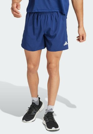 Homme portant un short de sport bleu, des chaussures de course noires et des chaussettes grises, debout devant un fond clair uni.