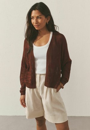 Žena má na sebe bordový pletený kardigán cez biely tielko a béžové plisované šortky, stojí s rukami v kapsách pri jednoduchom múre.