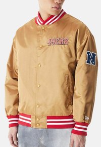 Giacca bomber in satin oro con colletto e polsini a strisce rosse e bianche, con "49ERS" ricamato in rosso e una patch "N" sulla manica.