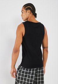Débardeur noir sans manches à coupe ajustée, fabriqué en matériel côtelé. Le modèle porte un short à carreaux noir et blanc avec une coupe ample.