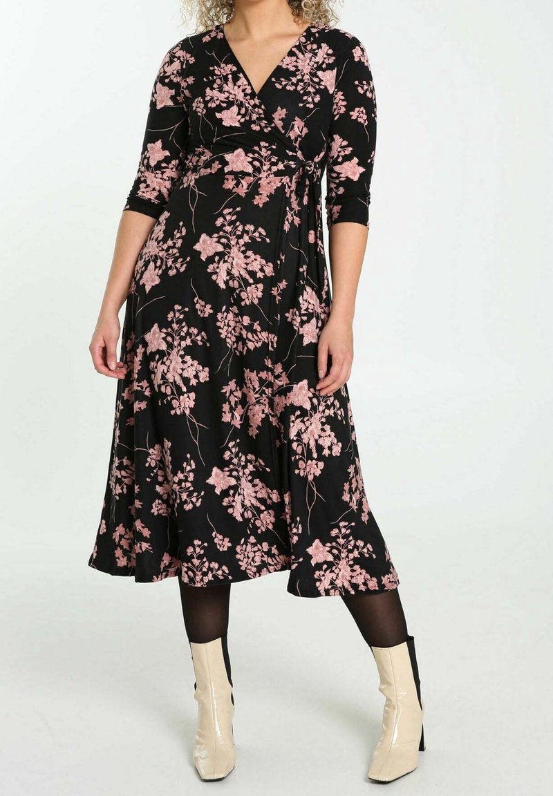 Femme portant une robe portefeuille midi noire avec un motif floral rose, manches trois-quarts, collants noirs et bottines crème.