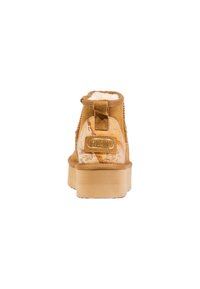 Bota de tobillo de ante marrón con un forro de felpa, un acento de logo dorado y una suela de plataforma beige texturizada que presenta un patrón de mapa.