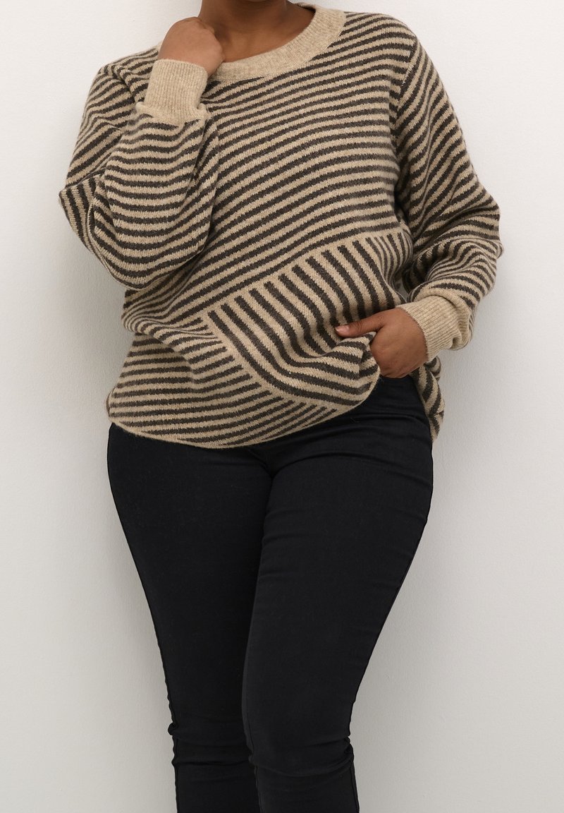 Pull en rayures beige et marron foncé à coupe ample, avec des poignets côtelés et un col rond, associé à un pantalon noir ajusté.