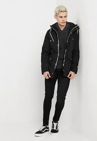 Casaco de penas preto com capuz, com fechos e botões prateados, usado sobre um suéter cinza e combinado com jeans pretos justo e ténis.