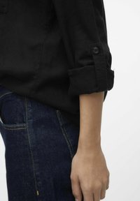 Camicia di lino nera con maniche arrotolate, caratterizzata da un singolo bottone nero. Indossata sopra jeans blu scuro con impunture dorate a contrasto.