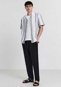 Uomo che indossa una camicia bianca a maniche corte con un motivo sopra una maglia bianca, pantaloni neri larghi e sandali neri, in piedi contro uno sfondo bianco.
