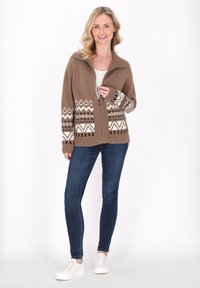 Bruine ritscardigan met geometrische witte en zwarte patronen op de mouwen, gedragen over een licht topje, in combinatie met donkere jeans en witte sneakers.