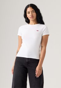 Bílý krátký rukáv tričko vyrobené z bavlny. Má malé červené logo Levi's na levém hrudníku. V kombinaci s černými širokými džíny.