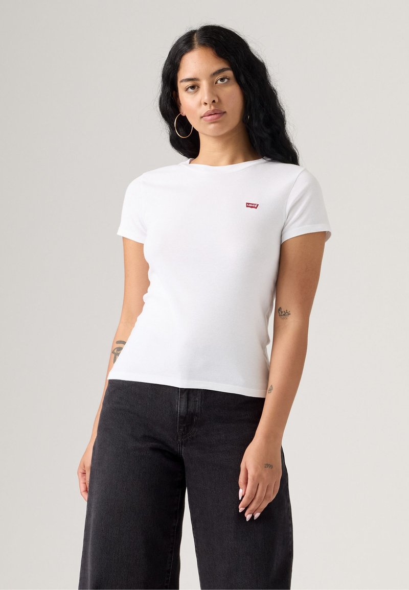 Bílý krátký rukáv tričko vyrobené z bavlny. Má malé červené logo Levi's na levém hrudníku. V kombinaci s černými širokými džíny.