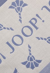 Lys beige stof med et gentaget mønster af blå blomstermotiver og ordet "JOOP!" i store bogstaver, blandet tekstur.
