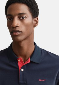 Námornícka polokošeľa s červeným golierom; dvojgombíkový plastrón s bielymi gombíkmi; vyšívané logo v červenej farbe na hrudi. Jemná textúra látky.