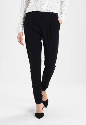 Vrouw draagt zwarte slim-fit broek, witte blouse met stippen en zwarte puntige hakken, staat met één hand in haar zak.