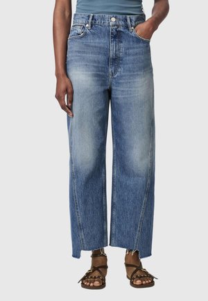 Persoon die versleten blauwe wijde jeans draagt met onafgewerkte zomen en bruine sandalen met riempjes, staand met één hand in de zak tegen een grijze achtergrond.