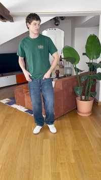 Zaļš kokvilnas T-krekls ar logotipu, zili džinsu bikses un baltas čības. Istabā ir brūns dīvāns un liels podiņš ar augu. Koka grīda.