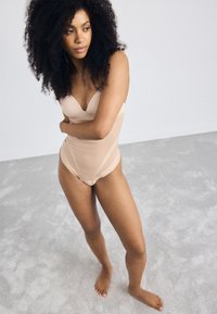 Beige body met v-halslijn, naadloos ontwerp en een gladde textuur. Het model heeft krullend haar en staat naaktvoets op een lichtgrijze ondergrond.