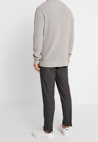 Mann in einem hellgrauen Strickpullover, dunkelgrauen Maßanzughose und weißen Sneakers vor einem schlichten weißen Hintergrund.