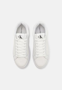 Sneakers bianchi con tomaia sintetica liscia, punta rotonda e lacci piatti. Presentano il logo nero "cK" sulla linguetta e il nome del marchio all'interno.