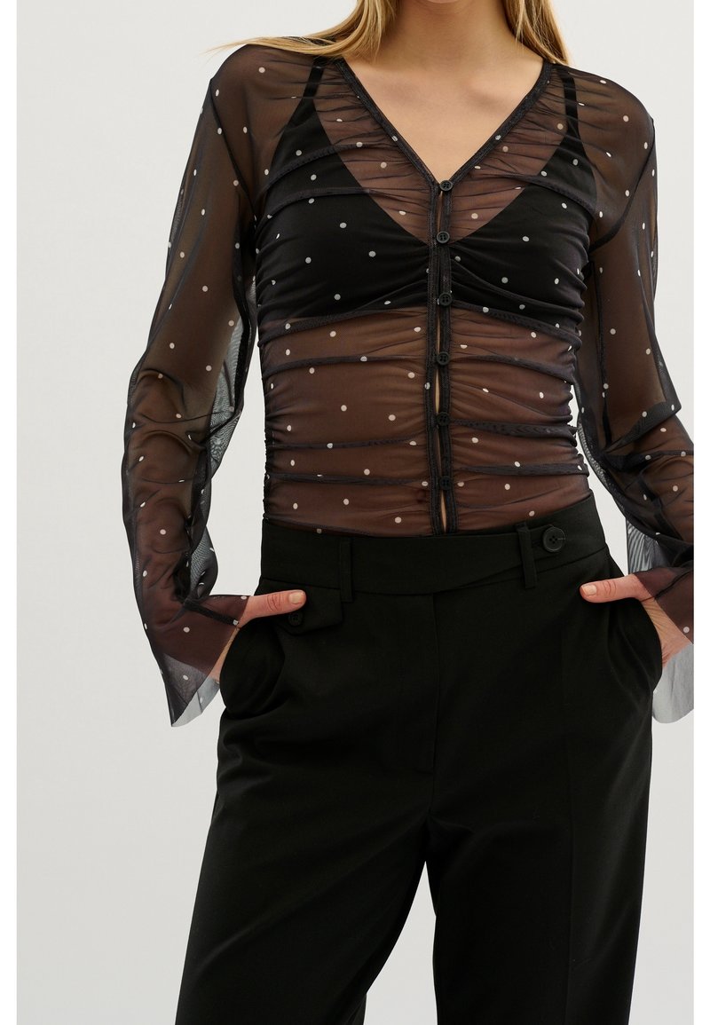 Svart genomskinlig blus med prickar, V-ringning, långa puffärmar och knappstängning. Kombination med högmidjade svarta byxor.