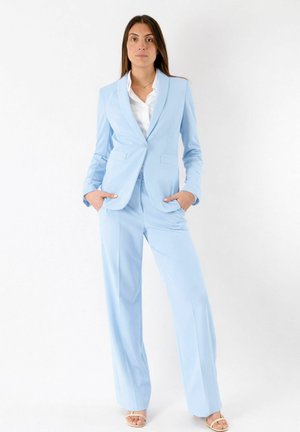 Donna in piedi con le mani nelle tasche, indossa un blazer azzurro chiaro e pantaloni a gamba larga coordinati su una camicia bianca e sandali.