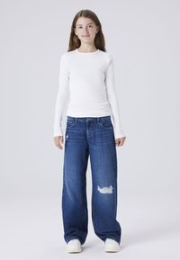 Camisa blanca de manga larga y jeans azules de gran tamaño con un parche desgastado en la pierna izquierda. Combinado con zapatillas blancas, sobre un fondo liso.