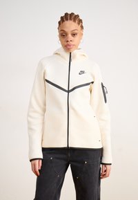Corta-vento Nike branco com capuz, fecho éclair preto e detalhes em preto, apresentando um design de chevron no peito e um bolso com fecho éclair na manga. Textura suave.