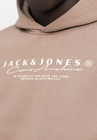 Felpa in cotone marrone con brandizzazione in testo bianco, che include "JACK & JONES" e "Core Design Department", con una superficie morbida e texturizzata.