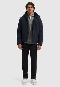 Donkerblauwe pufferjack met een capuchon, ritssluiting en zijzakken, gecombineerd met een grijze trui en zwarte broek, gedragen met witte sneakers.