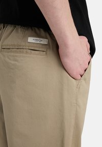 Khaki bukser med en glat tekstur, udstyret med en baglomme og et mærke syet nær taljen. Tætsiddende pasform.