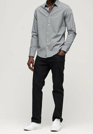 Uomo che indossa una camicia grigia con bottoni, pantaloni neri e scarpe da ginnastica bianche, in piedi con una mano in tasca davanti a uno sfondo semplice.