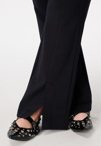 Zwarte, op maat gemaakte wijduitlopende broek met zijsplitten, gecombineerd met platte zwarte schoenen met metalen studs en een gesp.