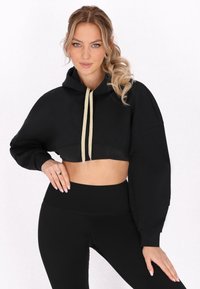 Felpa nera corta con cappuccio, coulisse beige in contrasto e maniche a sbuffo. Abbinata a leggings neri a vita alta.