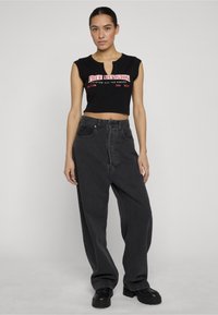 Naine musta lühikese varrukata toppi, millel on punane-valge tekst, lahtiste tumedate hallide kõrge vöökohaga teksade ja mustade massiivsete jalanõudega tavalise tausta ees.