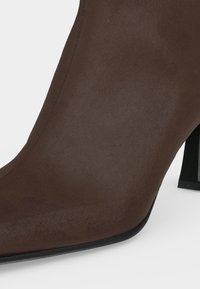 Bottine en daim marron à bout pointu avec talon bloc noir sur fond blanc.