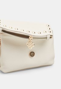Borsa in pelle color crema con borchie dorate lungo la parte superiore, chiusura con cerniera e un charm decorativo dorato. Dettaglio della superficie strutturata.