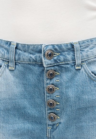Φωτεινό γαλάζιο τζιν με πέντε διακοσμητικά μεταλλικά κουμπιά και εμφανείς τρύπες κουμπιών, φορεμένο με λευκό ριμπ μπλουζάκι.