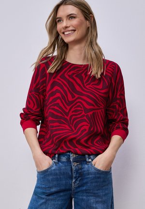 Młoda kobieta uśmiechająca się z rękami w kieszeniach, ubrana w czerwoną i ciemnofioletową koszulę w długi rękaw z wzorem oraz niebieskie dżinsy na jednolitym tle.