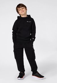 Ragazzo in piedi con le mani in tasca, che indossa un hoodie nero Champion, pantaloni cargo e sneakers nere con suola bianca e dettagli arancioni.