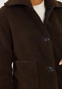 Bruine fleece jas met een gestructureerde oppervlakte, grote kraag en zwarte knoopsluitingen. Heeft een zijzak en contrasterende afwerking.