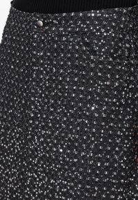 Jupe noire taille haute avec de petites paillettes argentées couvrant le tissu et une poche visible ainsi qu’un bouton près de la ceinture.