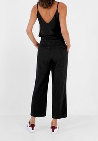 Femme portant une combinaison noire sans manches avec un pantalon large et des baskets blanches, debout avec les mains dans les poches, de dos.