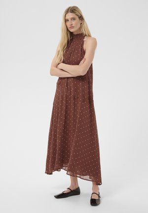Femme aux longs cheveux blonds portant une robe marron sans manches à pois blancs et des chaussures Mary Jane noires, debout les bras croisés.