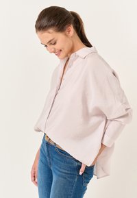 Jasnoróżowa, oversize koszula z kołnierzykiem, zapinana na guziki, z długim rękawem. Noszona na niebieskie dżinsy, które mają luźny krój i miękką teksturę.
