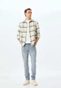 Camisa de franela a cuadros en crema y gris con acentos negros, camiseta blanca debajo, jeans de mezclilla azul claros y zapatillas beige.