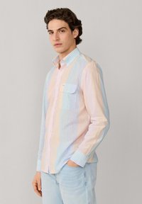 Jeune homme portant une chemise à rayures verticales pastel claires et un jean bleu clair, debout avec une main dans la poche sur un fond uni.