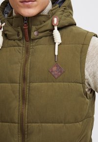Gilet matelassé vert olive avec capuche, doté d'un zip à l'avant, d'un patch en cuir avec logo et de cordons blancs. Tissu texturé et matelassé.
