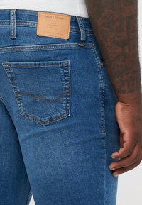 Primer plano de una persona que lleva vaqueros azules de Jack & Jones con costura en el bolsillo trasero, una etiqueta de cuero de la marca y un brazo tatuado visible.