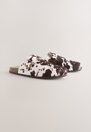 Mule s uzorkom smeđih i bijelih krava s ravnim potplatom, kožnim gornjim dijelom i bočnim remenom s kopčom. Imaju udobnu unutarnju podlogu.