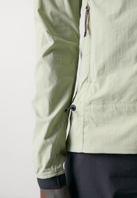 Veste vert clair en tissu texturé. Elle possède un cordon de serrage à l'ourlet et des poignets élastiques avec des accents en mesh noir. Poche zippée visible.
