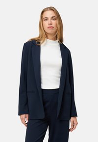 Blazer bleu marine en tissu texturé, doté d'une silhouette décontractée, de deux poches à l'avant et d'une fermeture à un seul bouton. Porté sur un haut blanc.