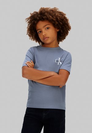 Copil cu păr creț purtând un tricou albastru Calvin Klein, cu brațele încrucișate, privind cu încredere către cameră, pe un fundal gri simplu.